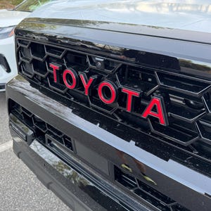 Puede incluir: Primer plano de una parrilla de coche negra con el logotipo "TOYOTA" rojo. La parrilla tiene un patrón de panal. Un coche blanco es parcialmente visible en el fondo.