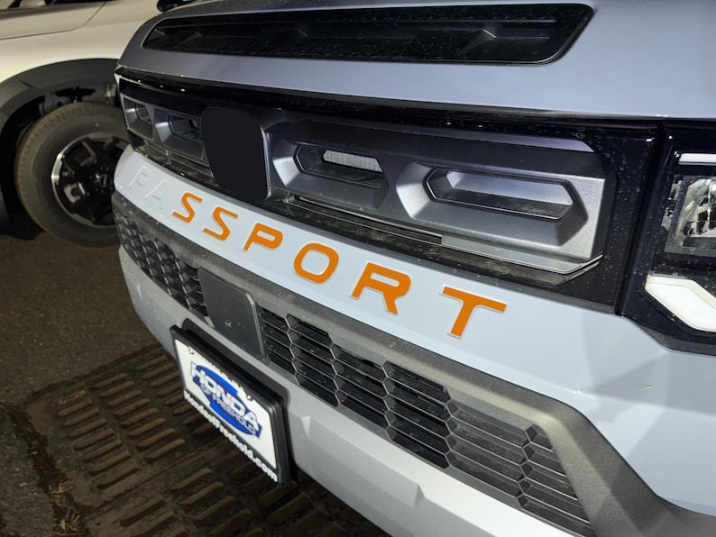 May include: A gray car with a black grille and the word "SPORT" in orange letters on the front bumper.