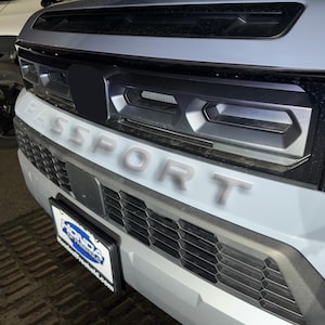 May include: A gray SUV with a black grille and the word "SPORT" written in gray letters across the front. The vehicle has a black license plate frame with the word "HONDA" printed on it.