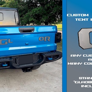 Peut inclure: Un camion bleu avec un badge de texte personnalisé sur le hayon. Le badge est argenté avec des lettres noires et dit "G". Le camion a des badges "GL" et "OR" sur le hayon. Le texte sur le côté droit de l'image dit "CUSTOM TAILGATE TEXT BADGE", "ANY CUSTOM TEXT AND MANY COLOR COMBO", et "STANDARD "GLADXXXXR" TEXT INCLUDED".
