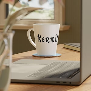 May include: A white ceramic mug with the word "Kermit" in black script. The mug sits on a light blue coaster on a wooden surface. A laptop and a window are in the background.