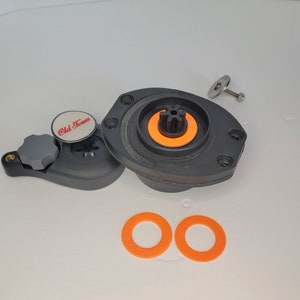 Könnte beinhalten: Eine schwarze und graue Kajakpumpe mit einem weißen Etikett mit der Aufschrift "Old Town". Die Pumpe hat einen schwarzen Knopf und einen grauen Griff. Auf einer weißen Oberfläche liegen zwei orangefarbene Gummringe.