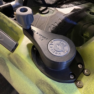 May include: A close-up of a black and gray kayak pedal drive system. The system has a gray handle and a black base with the text "PDL X-SPORT 3.5". The kayak is painted in shades of green and yellow.