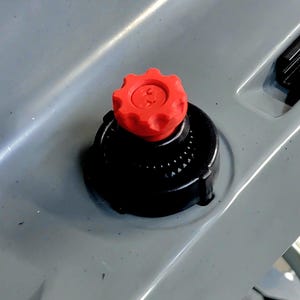 May include: Close-up of a red, gear-shaped knob atop a black, circular base. The knob is centered on a gray surface with a recessed area and a black, rectangular component visible.