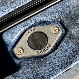May include: Close-up of a gray metal fitting with the word "TAX" and "HOBBY" inscribed. The fitting is secured with two screws and surrounds a black circular component. The background is a textured blue surface.