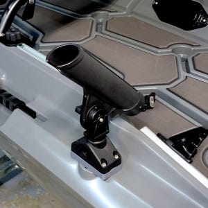 May include: A black fishing rod holder mounted on a gray kayak. The holder is cylindrical and angled upwards, with a pivoting base. The kayak has a gray and brown patterned deck.
