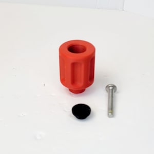 Könnte beinhalten: Ein roter Kunststoffknopf mit einer Schraube und einer schwarzen Kunststoffkappe. Der Knopf hat eine zylindrische Form mit einem gerippten Design.
