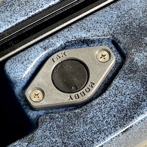 May include: Close-up of a gray metal fitting with the word "TAX" and "HOBBY" inscribed. The fitting is secured with two screws and surrounds a black circular component. The background is a textured blue surface.