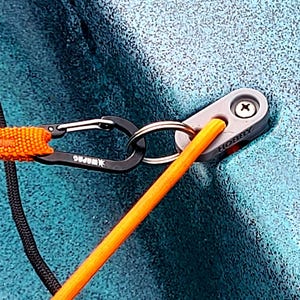 May include: Close-up of a kayak accessory. A gray metal fitting is secured with a screw, labeled "HOBBY". An orange strap connects to a black carabiner, attached to the fitting with a metal ring. A black rope is also visible.