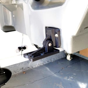 May include: A black transducer mount attached to a gray kayak. The mount is made of plastic and has a hinged design. The text "VAP ROLLY" is visible on the mount. The kayak is on a concrete surface.