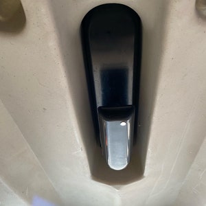 Op de afbeelding: Een close-up van het interieur van een boot, met een zwarte, langwerpige handgreep met een rechthoekig, verchroomd uiteinde. De handgreep is geplaatst tegen een lichtgekleurd, getextureerd oppervlak, met een gebogen ontwerp.