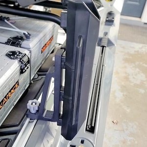 May include: A black plastic kayak accessory, mounted on a gray kayak. The accessory has a vertical storage compartment and is attached with a gray bracket and a knob. The kayak has the word "YAK" printed on it.