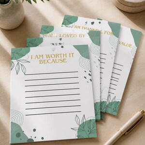 May include: A stack of white paper sheets with green abstract botanical illustrations and gold text. The top sheet reads I AM WORTH IT BECAUSE with black horizontal lines for writing. Other sheets feature text including I AM THANKFUL FOR and I AM LOVED BY.