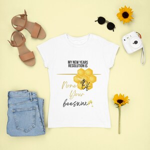 May include: White t-shirt with a graphic of a honeycomb and a bee. The text on the shirt reads "My New Years Resolution Is None Of Your Beeswax".