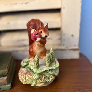 May include: A ceramic figurine of a squirrel holding a red fruit. The squirrel is brown and tan, perched on a base with green and tan foliage and small green trees. The figurine is set on a wooden surface.