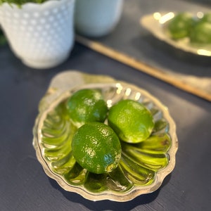 Könnte beinhalten: Drei grüne Limetten in einer silbernen, muschelförmigen Schale auf einer dunkelblauen Oberfläche.