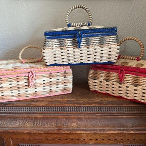 May include: Three vintage wicker baskets with lids and handles. The baskets are in different colors: pink, blue, and red. The baskets are sitting on a wooden surface.