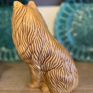 Könnte beinhalten: Eine Keramikfigur eines sitzenden Hundes, der den Kopf zurückdreht. Der Hund ist hellbraun mit braunen Felldetails. Die Figur ist mit strukturiertem Fell detailliert.