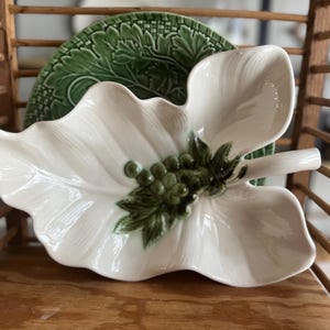 May include: White ceramic leaf-shaped serving dish with green grape and leaf accents. A green, textured plate is visible behind the leaf dish. The dish is on a wooden shelf.