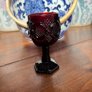 Puede incluir: Un pequeño cáliz de vidrio rojo oscuro con un diseño estampado. El cáliz tiene un tallo negro y una base cuadrada negra.