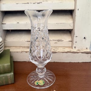 May include: A clear crystal vase with a diamond-cut pattern. The vase has a flared top, a slender body, and a round base. A small green and gold sticker is on the base. The vase is on a wooden surface.