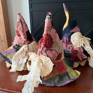 Puede incluir: Tres sombreros de bruja hechos a mano con tela de retazos en tonos rosa, burdeos y verde. Cada sombrero presenta un lazo de encaje decorativo y una base de mimbre. Los sombreros se exhiben sobre una superficie de madera.