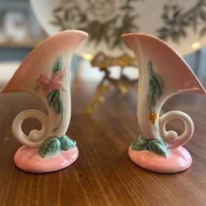 May include: Two small, pink and white ceramic vases with floral designs. The vases are shaped like trumpets and have a pink base.