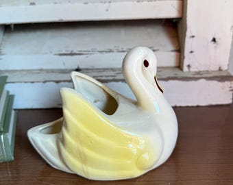 May include: A ceramic swan figurine with a white body and yellow wings. The swan has a long neck, a red eye, and a yellow beak. The figurine is sitting on a wooden surface.