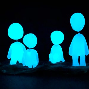 May include: Four glowing blue figures made of glass or ceramic stand on a dark surface. The figures are all different sizes and shapes, and they are all lit from within, creating a soft, ethereal glow.