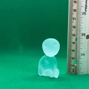 May include: A clear glass figurine of a person sitting with their legs crossed. The figurine is about 2 inches tall and is sitting next to a ruler.