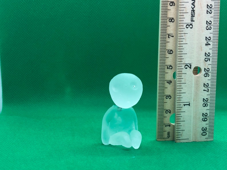 May include: A clear glass figurine of a person sitting with their legs crossed. The figurine is about 2 inches tall and is sitting next to a ruler.