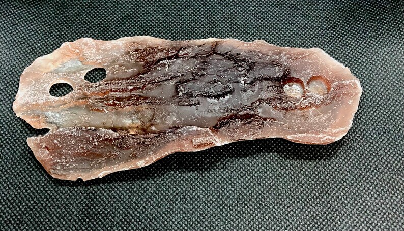 May include: A light brown, translucent, irregularly shaped piece of selenite with two holes drilled through it. The selenite has a rough, textured surface and a smooth, polished edge.