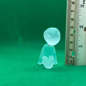 May include: A clear glass figurine of a person sitting with their legs crossed. The figurine is about 2 inches tall and is sitting on a green surface next to a ruler.