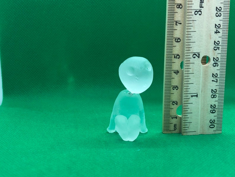 May include: A clear glass figurine of a person sitting with their legs crossed. The figurine is about 2 inches tall and is sitting on a green surface next to a ruler.