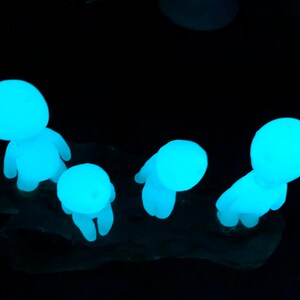 May include: Four small, glowing blue figurines in the shape of people standing on a dark surface. The figurines are made of a translucent material and are glowing in the dark.