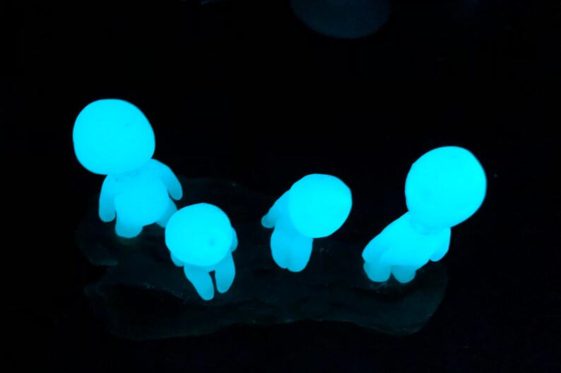 May include: Four small, glowing blue figurines in the shape of people standing on a dark surface. The figurines are made of a translucent material and are glowing in the dark.