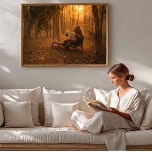 Può includere: Una stampa incorniciata raffigura un orso che legge un giornale su una panchina in una foresta illuminata dal sole. L'opera d'arte è sopra un divano color crema con cuscini bianchi. Una donna è seduta sul divano, leggendo un libro.