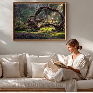 Può includere: Una stampa artistica incorniciata raffigura una scena di foresta con un grande albero ad arco e una volpe. L'opera d'arte è appesa sopra un divano color crema con cuscini. Una donna è seduta sul divano, leggendo un libro, con un abito chiaro.