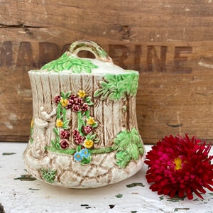 May include: A ceramic sugar bowl with a lid, decorated with a floral design and a tree trunk pattern. The bowl is white with green, brown, red, and yellow accents.