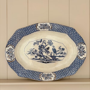 May include: An oval ceramic serving platter featuring a cream-coloured centre with a blue transferware illustration of a crane, trees, and floral motifs. The wide rim has a dark blue geometric pattern with four decorative floral cartouches.