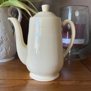 May include: Cream-coloured ceramic teapot with a lid and a curved spout. The teapot has a rounded base and a handle. The teapot is sitting on a wooden surface. A white vase and a glass candle holder are in the background.