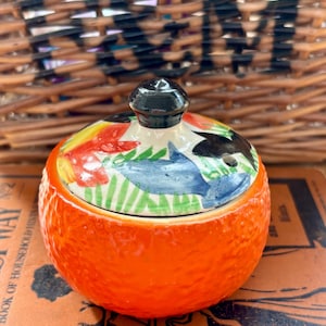 May include: A round ceramic sugar bowl with a textured, bright orange exterior resembling an orange peel. The lid features a hand-painted design with blue, green, red, and black shapes, topped with a small, smooth, black spherical handle.