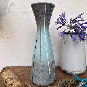 May include: A tall, slender ceramic vase featuring a gradient colour scheme that transitions from dark grey at the top to a light blue in the centre and back to a speckled grey at the base, accented with vertical brown and white painted stripes.