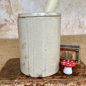 May include: A vintage stoneware crock with a cylindrical shape, holding a cream-coloured candle. A small red and white mushroom figurine and a matchbox are next to the crock. The items are on a book.