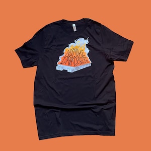 Puede incluir: Una camiseta negra de manga corta con un gráfico de un edificio envuelto en llamas naranjas y amarillas con humo azul. El texto "NO MORE DATACENTERS" está escrito en letras negras sobre el fuego, y se ve un pequeño logotipo en el edificio.