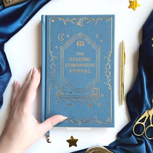 May include: A blue journal with gold foil detailing, titled "The Reading Companion Journal". The cover features a decorative design with a moon, stars, and floral elements. A gold pen and scissors are also visible.