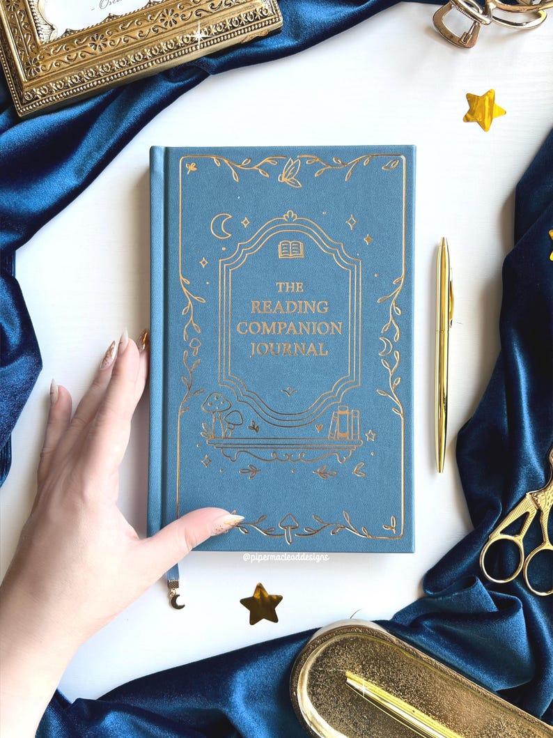 May include: A blue journal with gold foil detailing, titled "The Reading Companion Journal". The cover features a decorative design with a moon, stars, and floral elements. A gold pen and scissors are also visible.