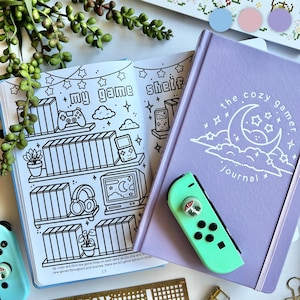 May include: A light purple journal titled the cozy gamer journal with a white moon and cloud graphic, paired with a mint green game controller featuring a mushroom thumb grip. An open colouring book page shows a shelf illustration with game consoles.