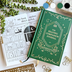 May include: A green hardback journal with gold detailing, labelled "The Reading Companion Journal". The journal is open to a black and white coloring page with a bookshelf illustration. Gold binder clips and a stencil are also visible.
