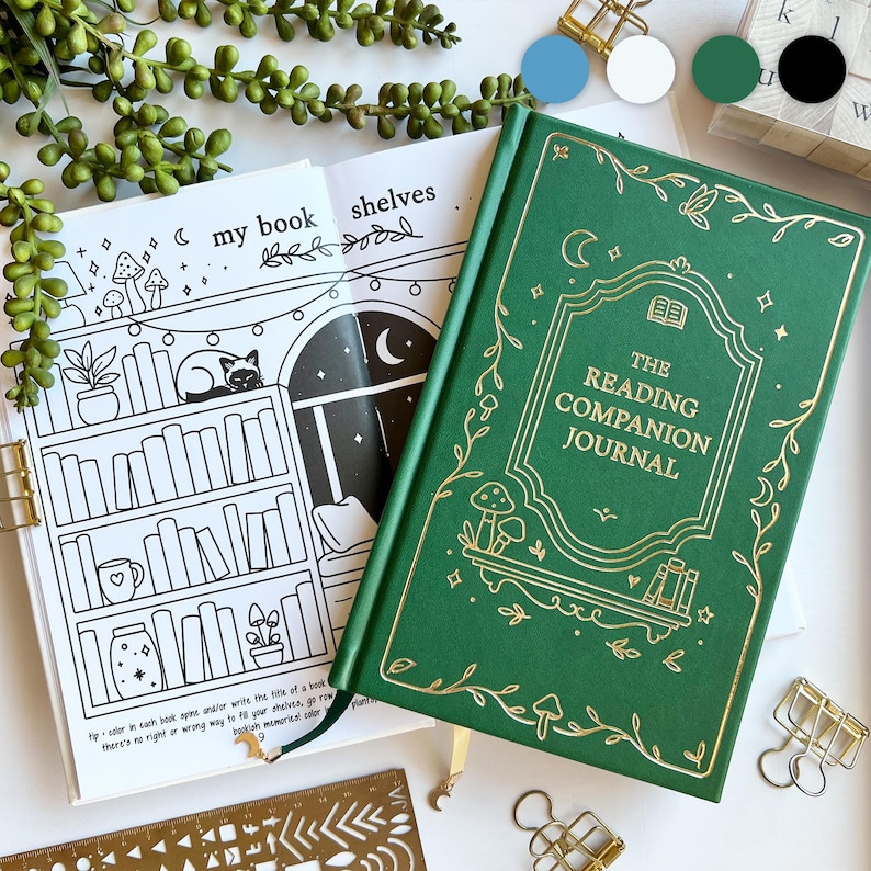 May include: A green hardback journal with gold detailing, labelled "The Reading Companion Journal". The journal is open to a black and white coloring page with a bookshelf illustration. Gold binder clips and a stencil are also visible.